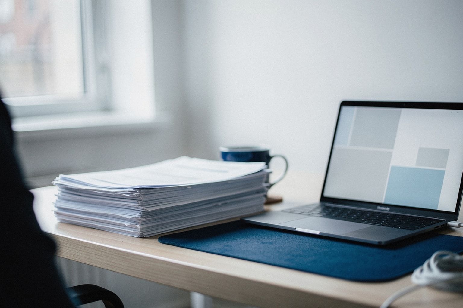 Dicker Stapel Papierformulare neben einem Laptop mit aufgeräumter Software-Oberfläche